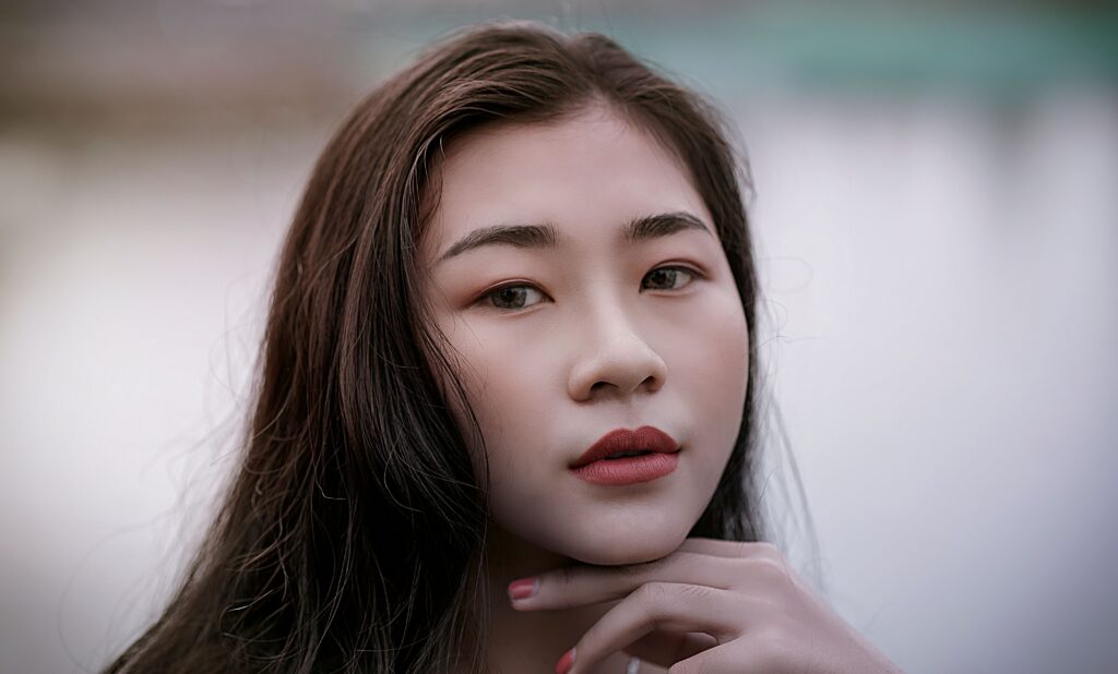 Close-up portrait of a young woman with long dark hair, pink lipstick, and hands gently resting under her chin, looking slightly to the side.