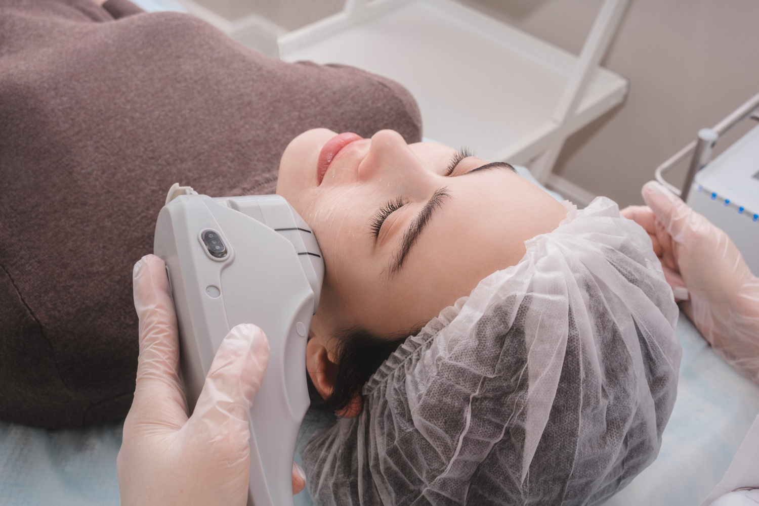 Woman lying with a hair cap during a cosmetic facial treatment; clinician uses a handheld device on her jaw.