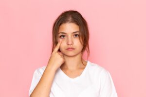 Young woman in a white shirt touching the area under her eye with her index finger against a pink background.