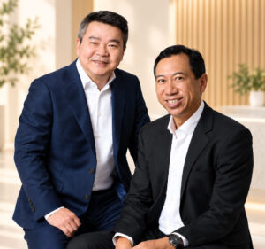 Two professionally dressed men in suits pose together and smile in a bright lobby setting.