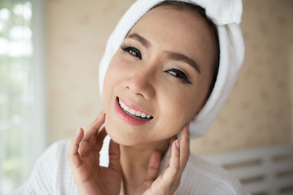 Smiling woman with a white towel wrapped around her head, close-up portrait indoors, wearing light makeup and pink lips